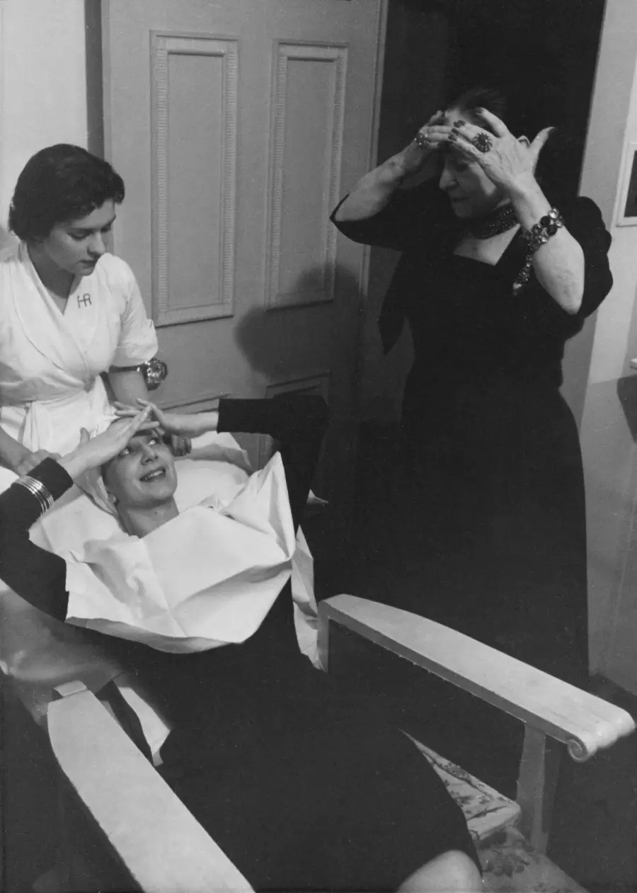 A woman sits in a chair with papers on her chest, touching her forehead, while another woman in black does the same standing nearby; a nurse or attendant stands by watching.
