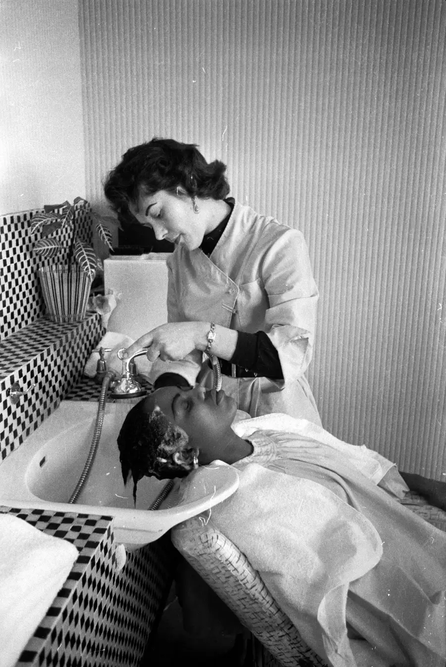 A hairstylist washes a woman’s hair at a salon sink. The stylist is focused, while the client reclines with eyes closed, wearing a protective cape. The salon features checkered tile and striped wallpaper.
