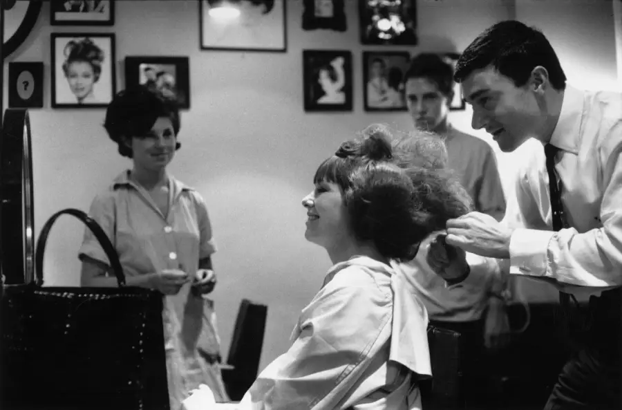 A hairstylist styles a smiling woman’s hair in a salon, while two people watch. Portraits and hairstyle photos hang on the wall in the background.