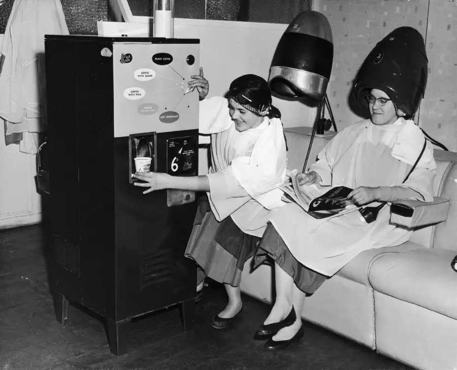 Two women at a hair salon; one uses a vending machine while the other sits under a large hair dryer, reading a magazine. Both wear salon smocks and vintage hair curlers in a black and white setting.