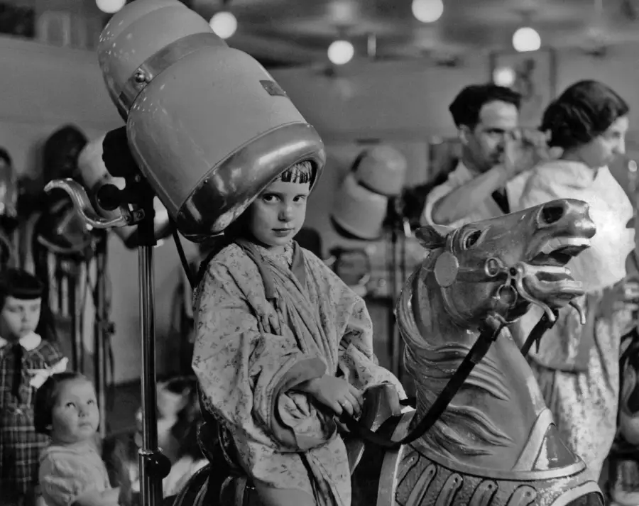 A young girl sits on a carousel horse while wearing a vintage hair dryer at a busy salon. Other children and adults in gowns and hair dryers are visible in the background. The scene is in black and white.