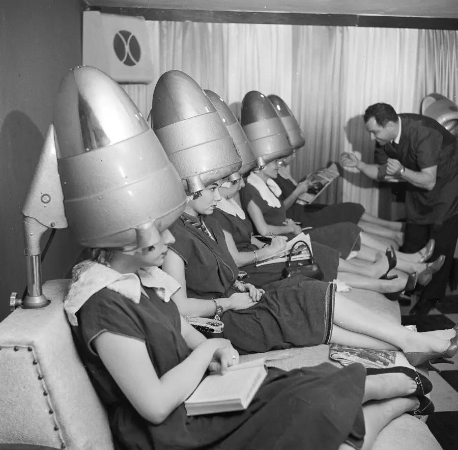 Four women sit under vintage hair dryers in a salon, wearing capes and reading. A stylist stands nearby, attending to one of the women. The photo is in black and white and has a retro, mid-20th-century feel.