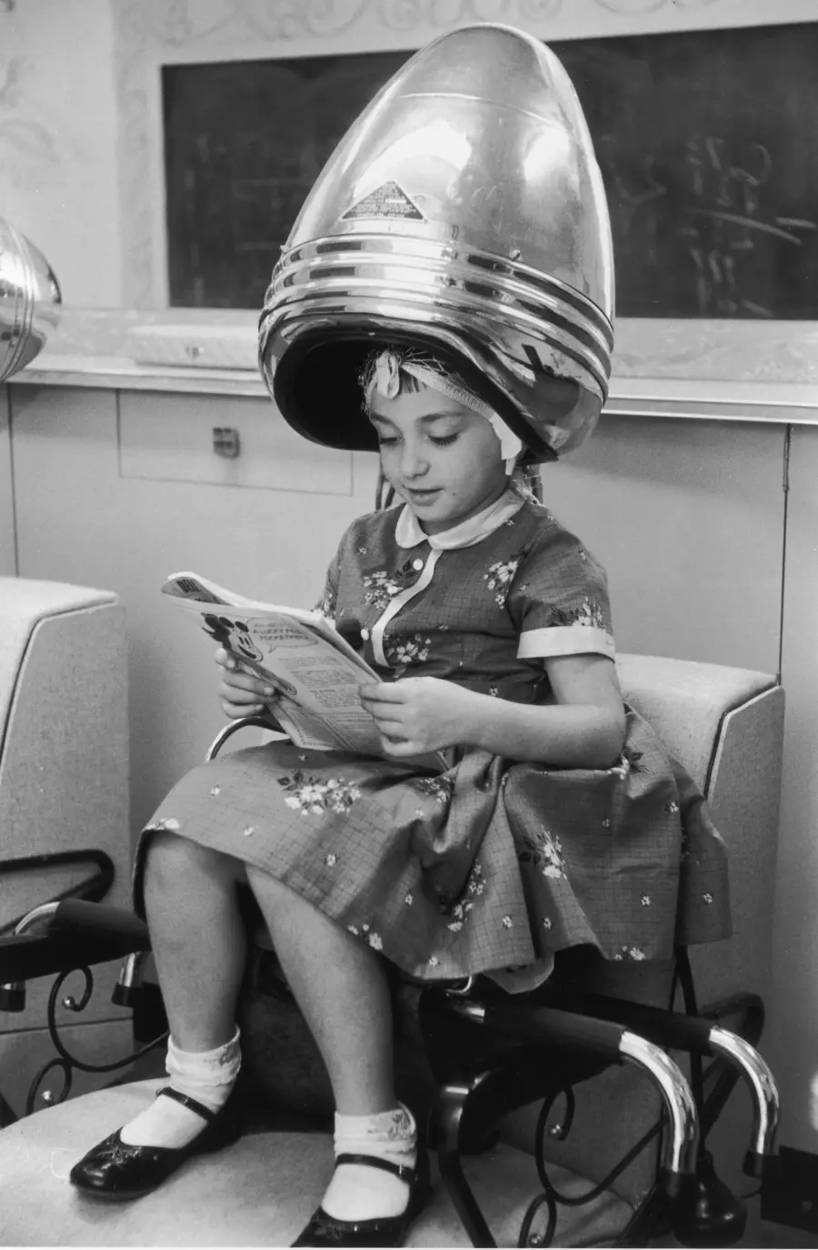 A young girl in a dress sits under a vintage hair dryer at a salon, reading a magazine and wearing socks with Mary Jane shoes.