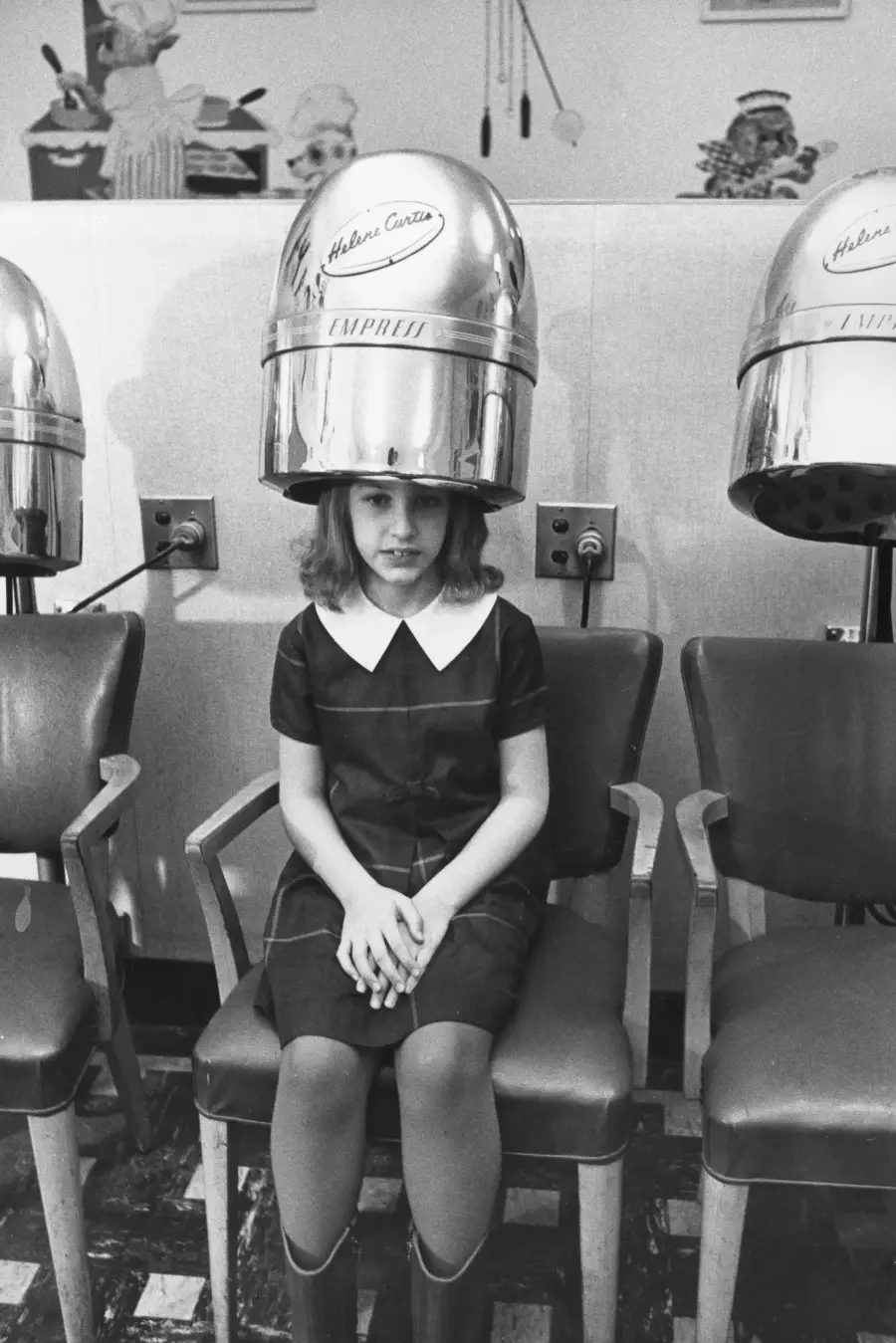 A young girl sits under a vintage hair dryer in a salon, wearing a dark dress with a white collar. She has her hands clasped in her lap and looks slightly nervous. Two empty salon chairs are beside her.