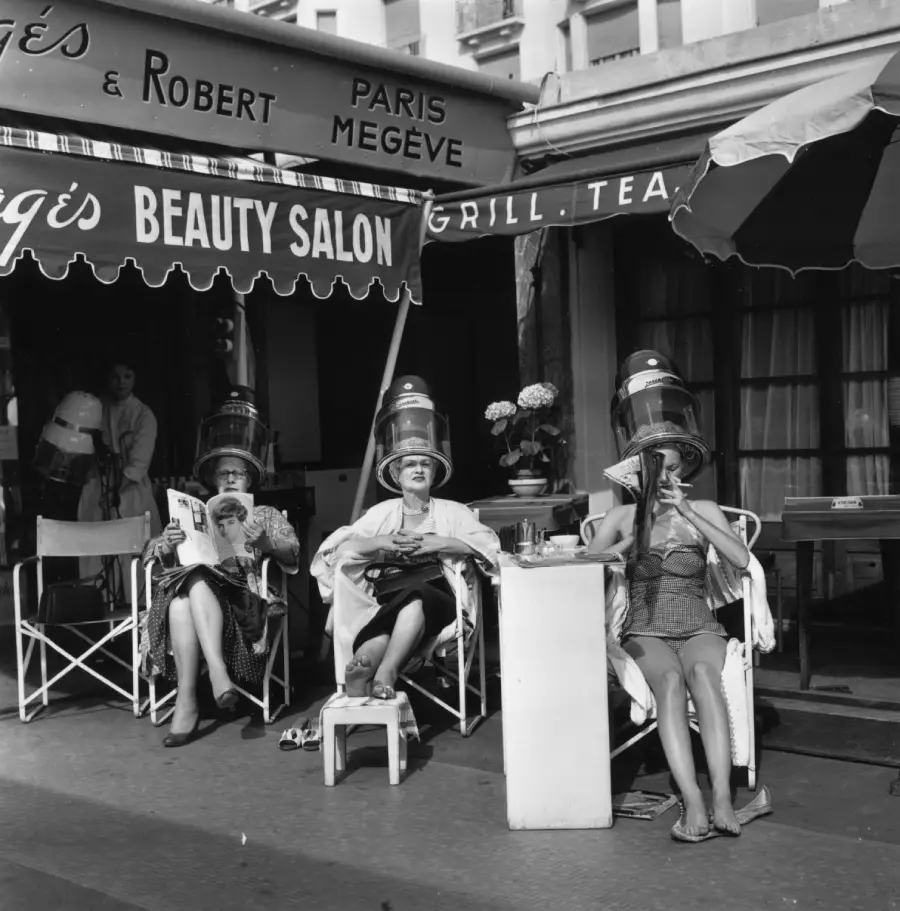Three women sit outside a beauty salon under vintage hair dryers, reading and relaxing. A salon worker stands in the doorway. The scene is lively, with salon and café awnings overhead and potted flowers nearby.