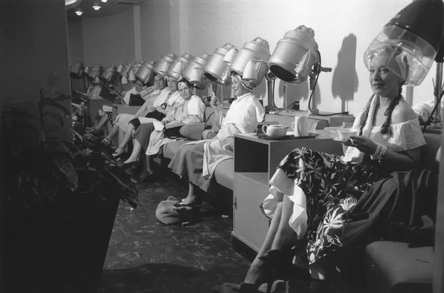 Black and white photo of women sitting in a row at a salon under vintage hair dryers, some drinking tea. They wear salon capes and have towels on their laps. The mood is relaxed and social.