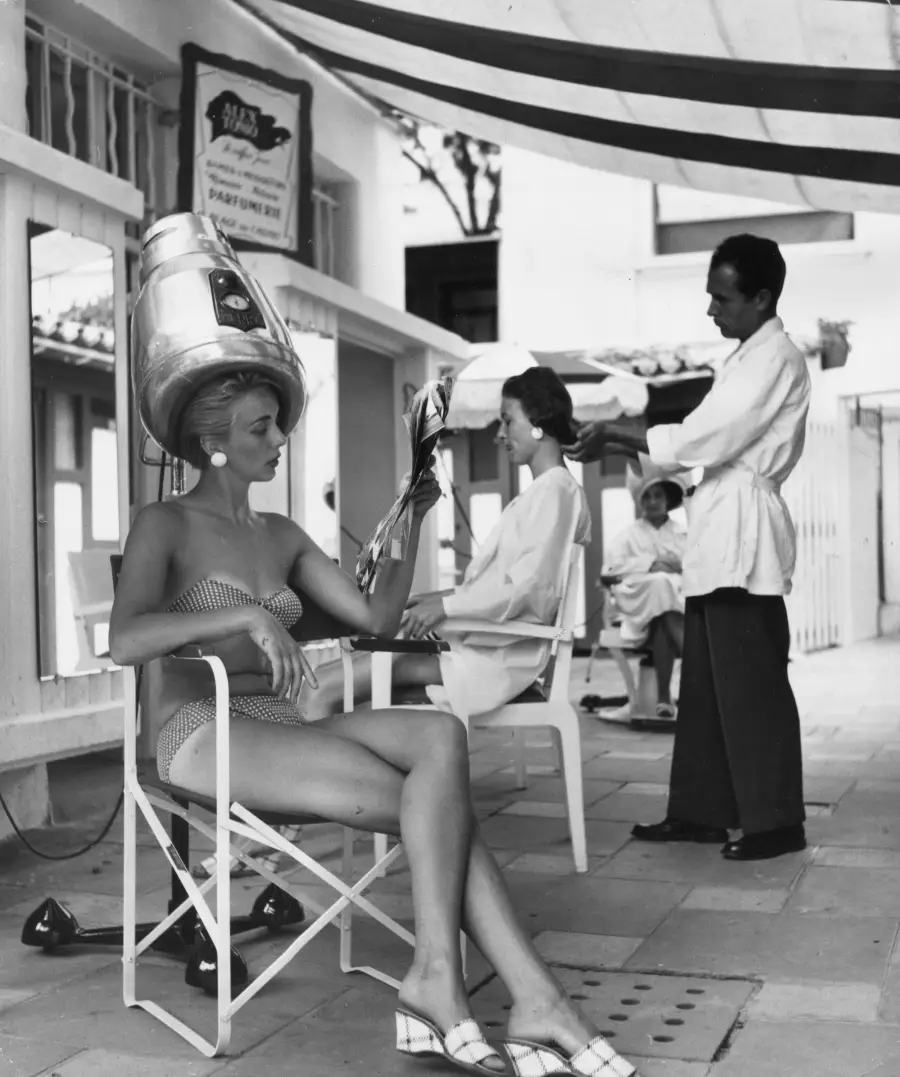 A woman in a swimsuit sits under a hair dryer, reading a magazine, while another woman gets her hair styled by a man in a white coat at an outdoor salon.