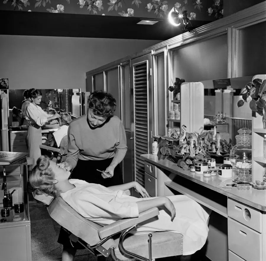 A woman reclines in a salon chair while another woman applies makeup at a vanity. The room has mirrors, shelves with beauty products, and floral wallpaper. Another client and stylist are visible in the background.