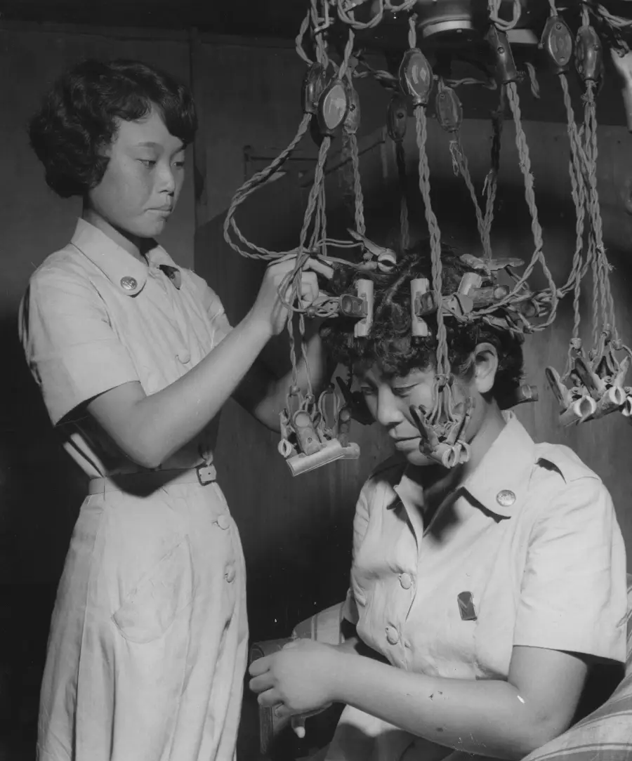 A woman adjusts a vintage perm machine on another woman’s head in a salon. Both wear uniforms. Numerous wires and rollers are attached to the seated woman’s hair.