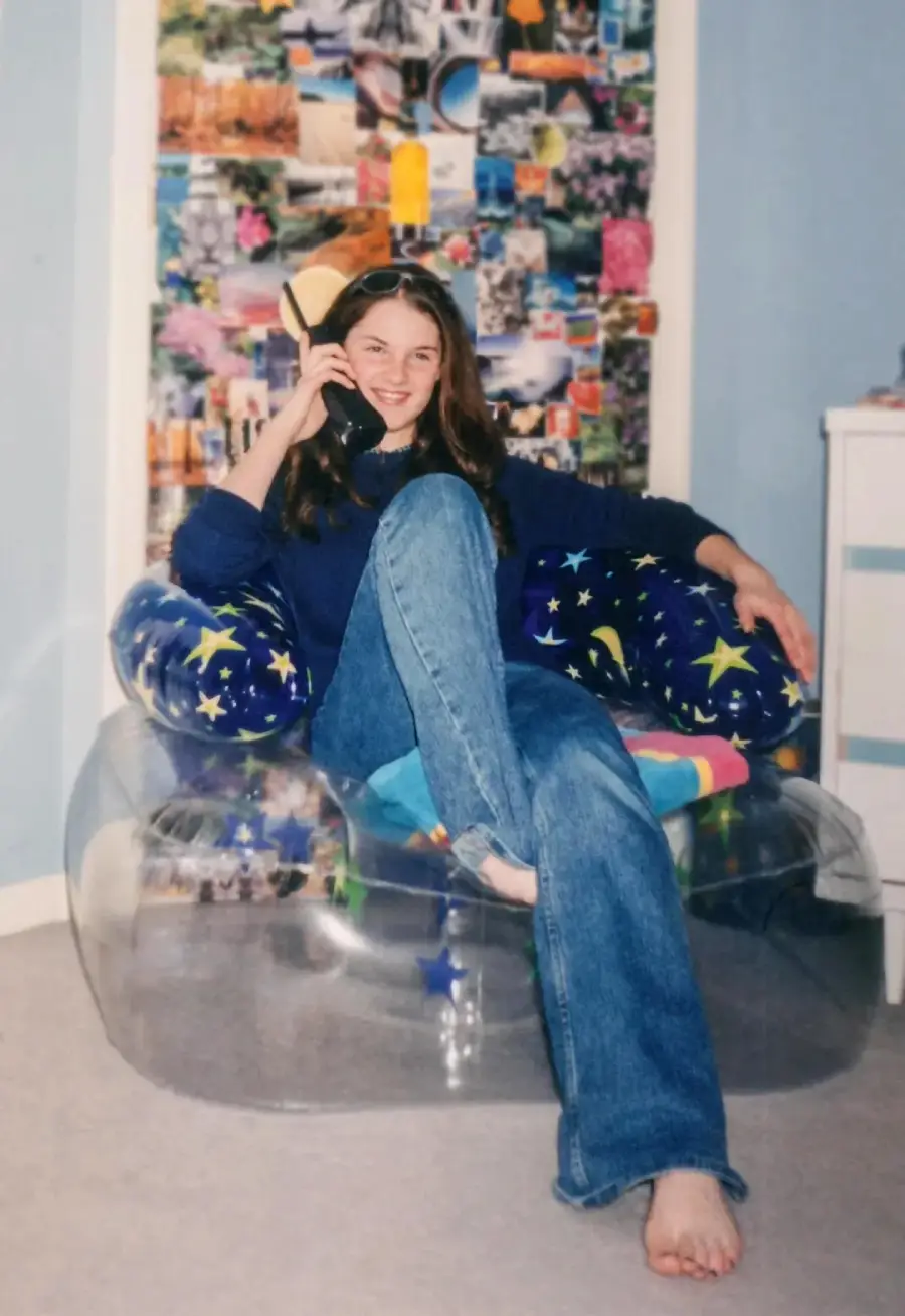 Teen girl sitting barefoot on a clear inflatable chair, talking on a corded phone. She wears jeans and a blue sweater. Behind her is a wall collage of colorful photos and a blue wall. A dresser is partly visible on the right.