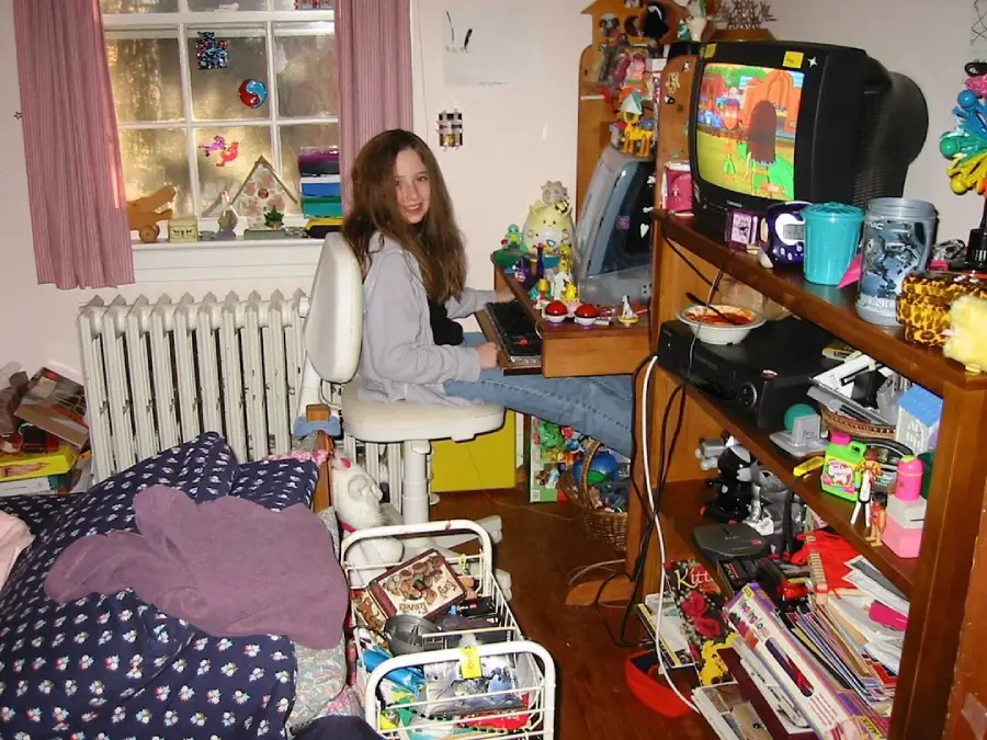A girl with long brown hair sits at a computer desk in a cluttered bedroom, smiling at the camera. The room has shelves with books, toys, and a TV displaying a colorful game. A messy bed is in the foreground.