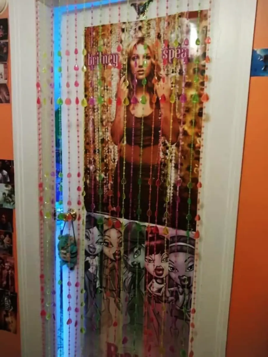 A door decorated with a Britney Spears poster, Bratz posters, and colorful beaded curtains. The room has orange walls with photos and blue light seen through the edge of the door.