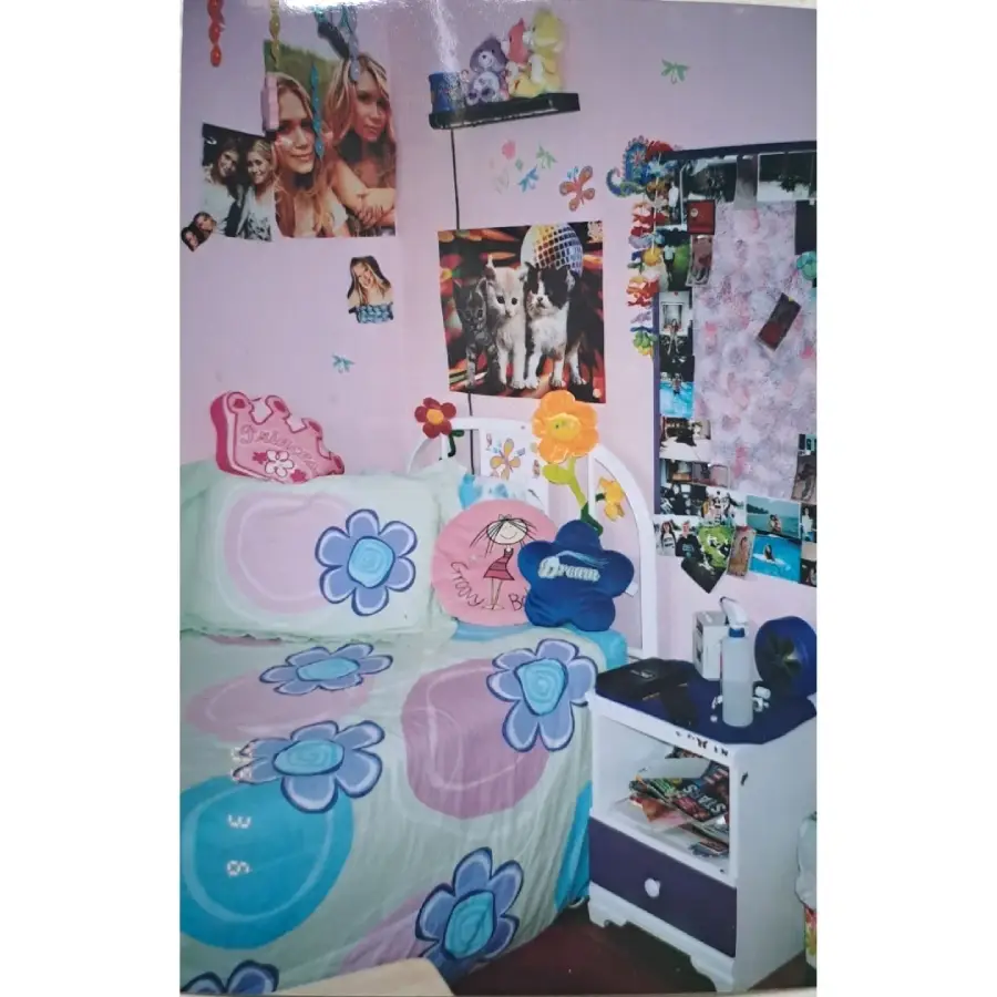 A pastel purple bedroom with a floral bedspread, plush toys, celebrity posters, and photos on the walls. A white nightstand holds a lamp, mug, and small items. The room feels colorful and personalized.