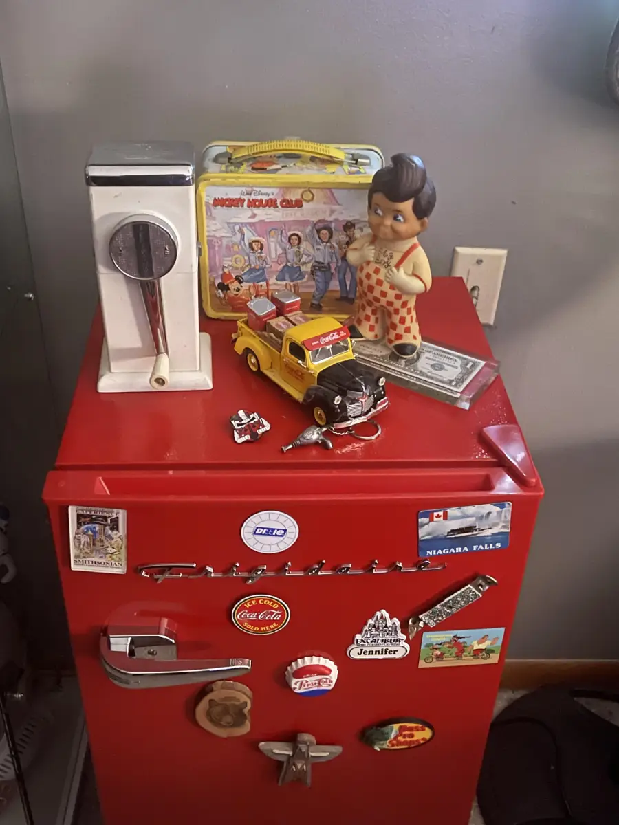 A red mini fridge with vintage magnets, a toy car, a dollar bill, a doll, an old lunchbox, and a white scale on top. The background includes a gray wall and an electrical outlet.
