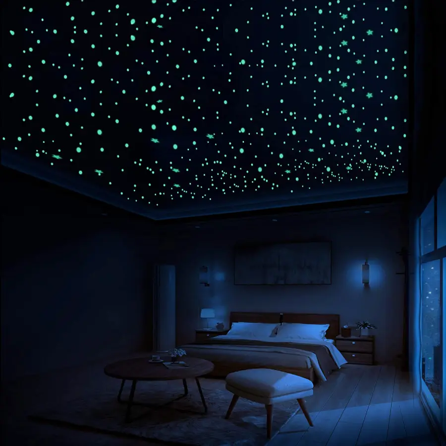 A dark bedroom with glow-in-the-dark stars on the ceiling, creating a starry night effect. There is a bed, nightstands, a round table, an ottoman, and a window showing the outdoors.