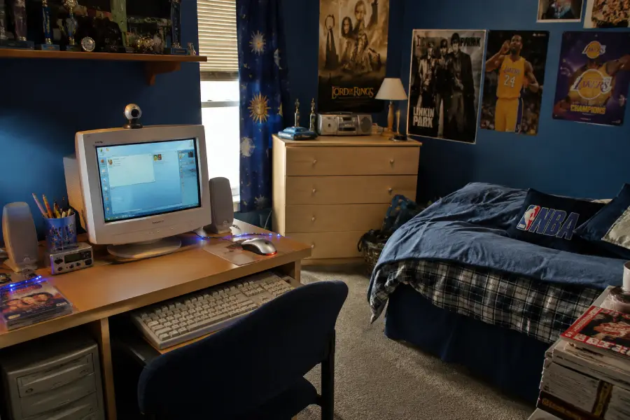 A blue-themed bedroom with a desk holding an old computer, shelves with trophies, a bed with plaid bedding, posters on the walls, and magazines stacked near the bed. Sunlight streams through a window with celestial curtains.