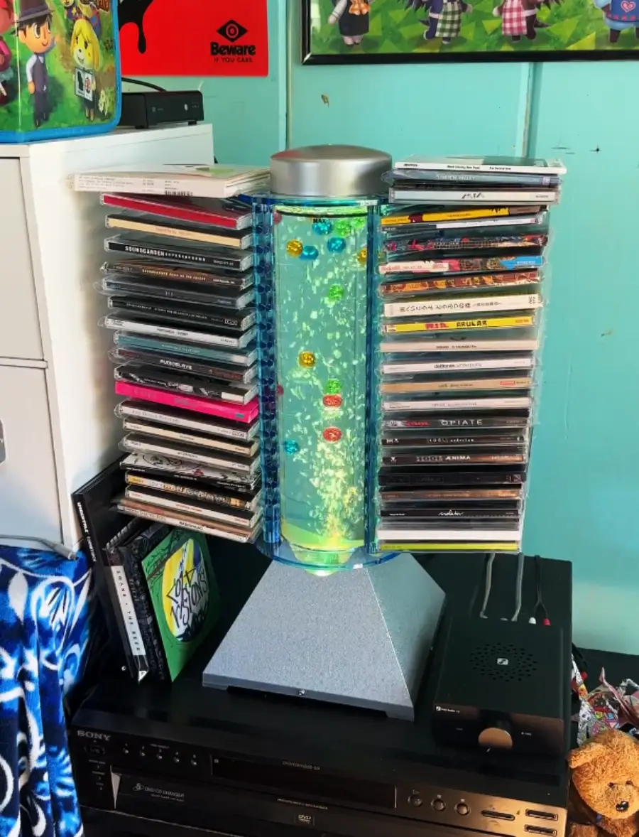 A lava lamp is used as a stand for storing CD cases on both sides. The setup is on top of electronic equipment in a room with blue-green walls and various posters and items around.