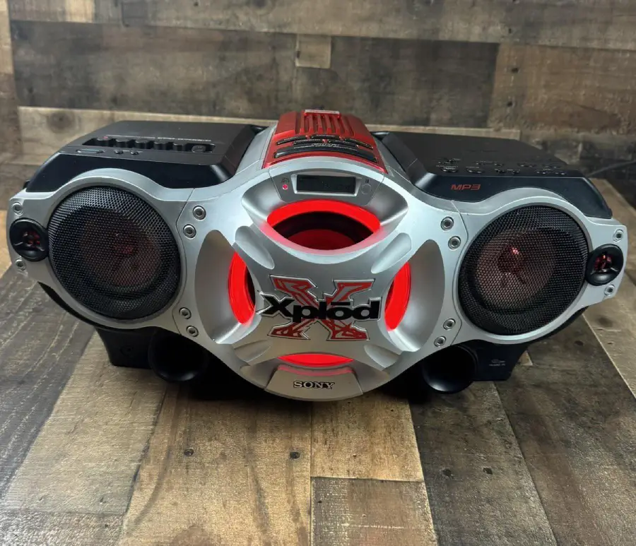 A Sony Xplod boombox with a silver and red design, black speakers on each side, and visible control buttons, placed on a wooden surface with a wooden wall background.