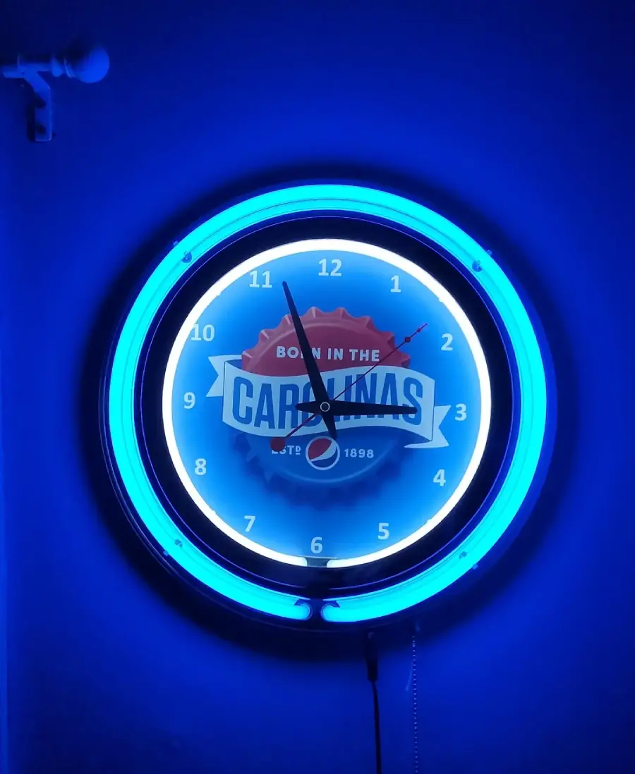 A neon blue wall clock with the words “Born in the Carolinas” on its face. The time shows 11:00. The clock is glowing brightly in the dark.