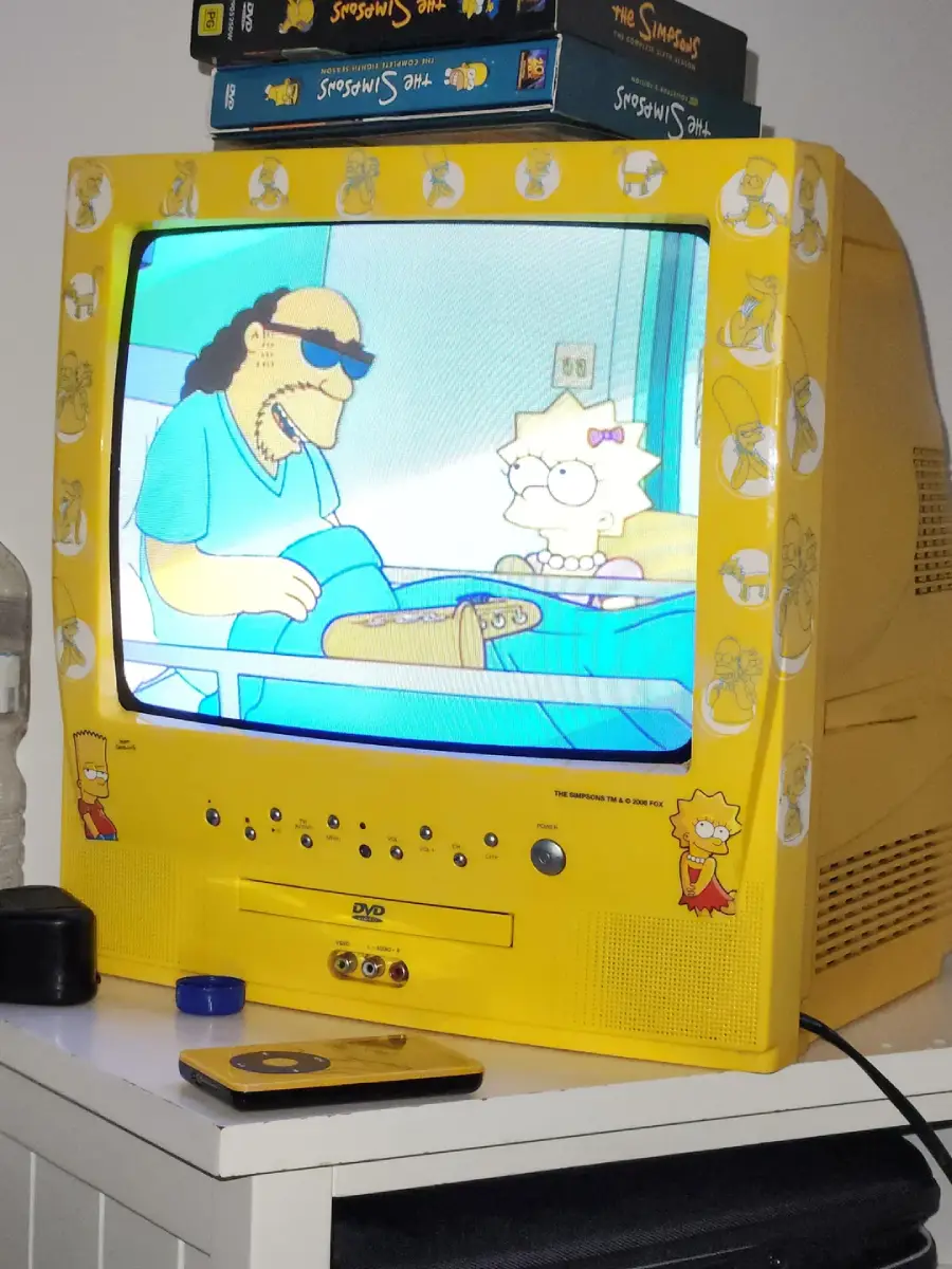 A yellow TV with Simpsons stickers shows a Simpsons episode. Simpsons DVD cases are stacked on top, and Simpsons DVD and remote are beside the TV on a white table.