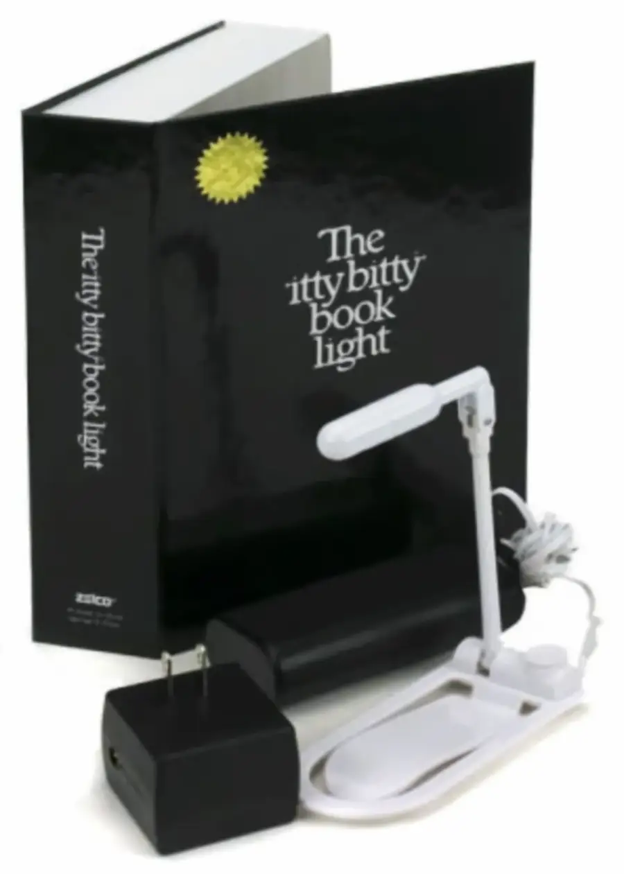 A box labeled “The itty bitty book light” stands behind a small, white, flexible book light, a power adapter, and a case, all arranged on a white surface.