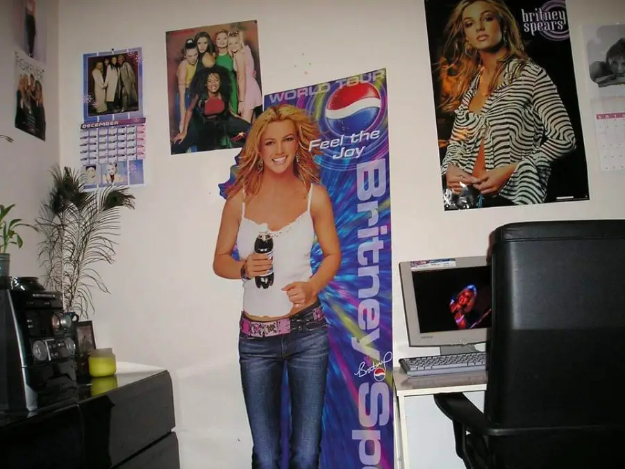 A room with Britney Spears posters on the wall, a cardboard cutout of her, a desk with a computer, a chair, and a stereo.