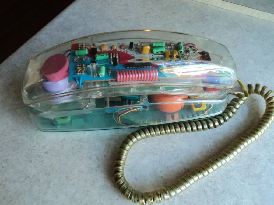 A clear plastic landline telephone showing colorful internal components, with a coiled cord attached, resting on a light-colored countertop.