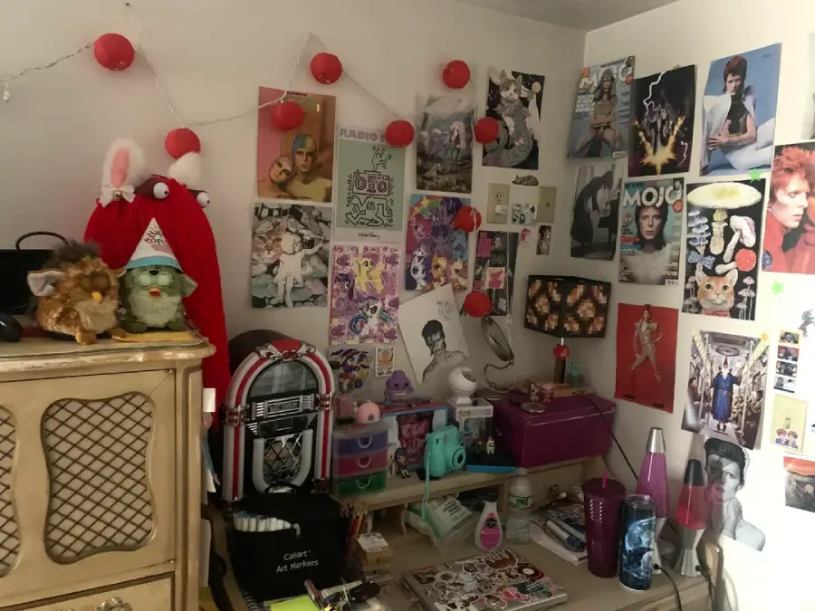 A cluttered desk with a lava lamp, jukebox, Furbies, art supplies, and various posters and photos covering the wall, featuring musicians, animals, and colorful illustrations.