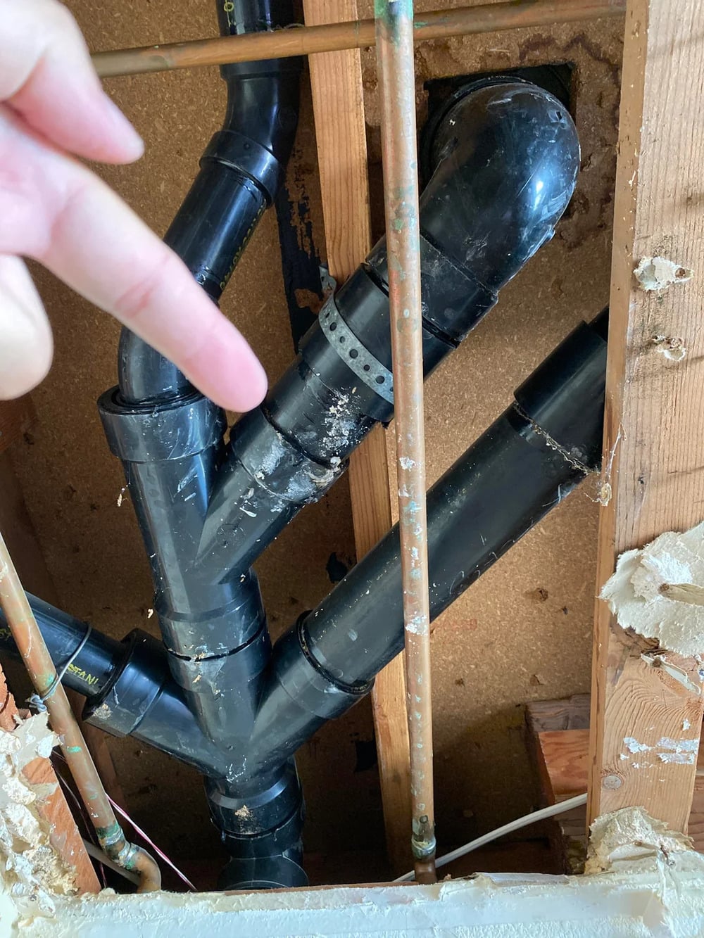 A hand points at exposed black plastic plumbing pipes and copper water lines inside a wall, showing plumbing joints and fittings with insulation and drywall partially removed.