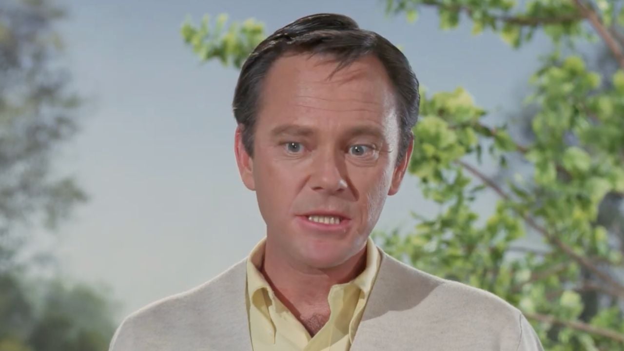 A man with short dark hair wearing a light yellow shirt and beige sweater stands outdoors with green trees and a blue sky in the background.