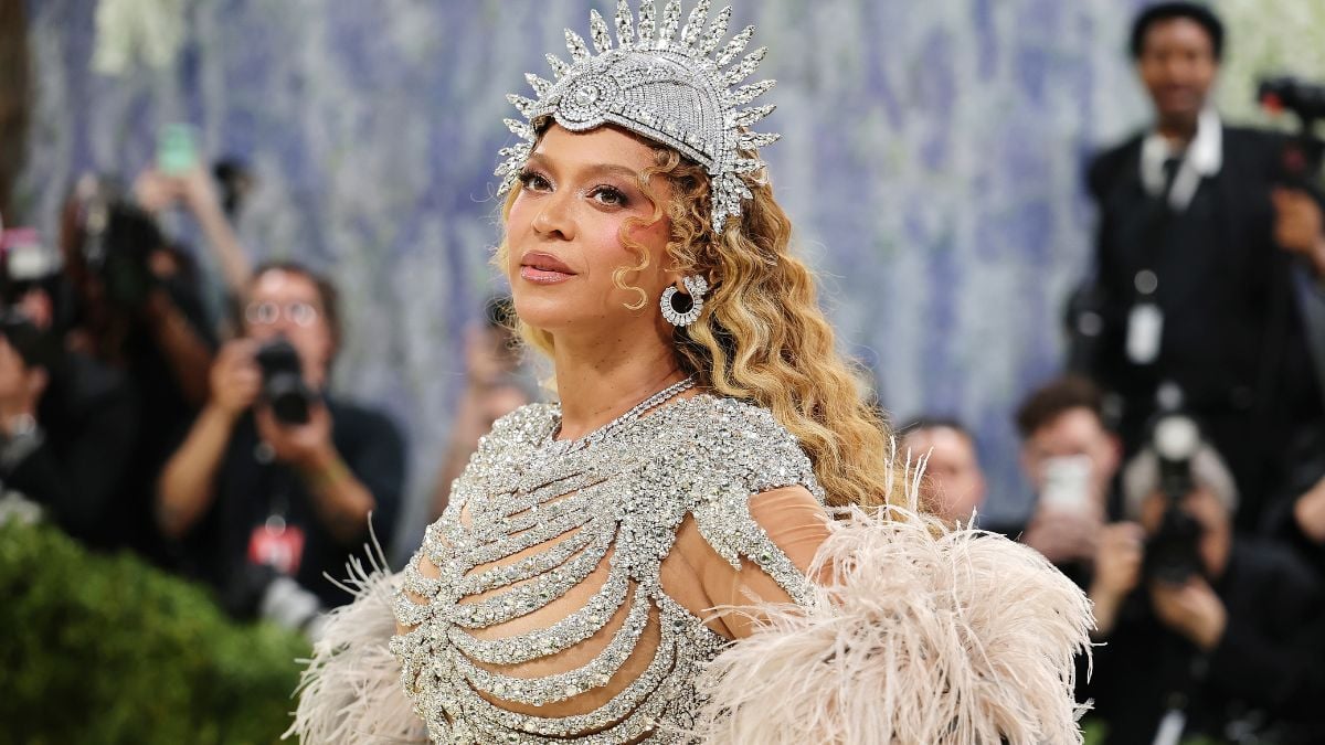 A woman in a sparkling silver gown with an elaborate headpiece poses on a green carpet, surrounded by photographers. She has long, curly hair and wears dramatic feathered sleeves.