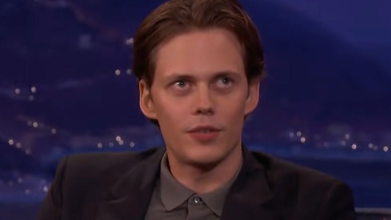 A man with light skin and brown hair, wearing a dark jacket and gray shirt, appears to be speaking during a television interview. The background is blurred with blue and purple tones.