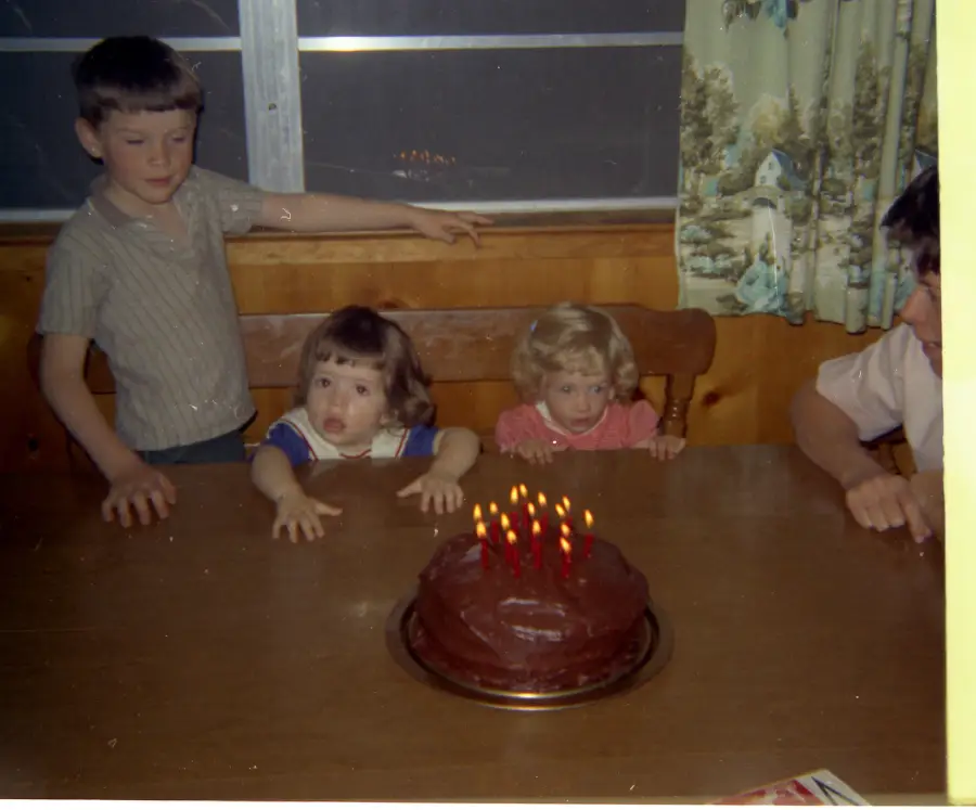 Three young children sit at a wooden table, looking at a chocolate birthday cake with six lit candles. An adult’s arm is visible at the right edge of the image. The scene appears to be indoors, near a window.