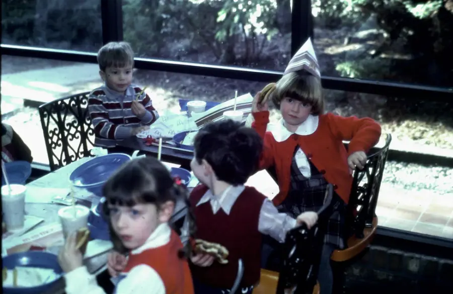 Four children sit at a table with food and drinks. One child wears a birthday hat and holds up a hot dog. The scene is indoors by large windows with greenery outside.
