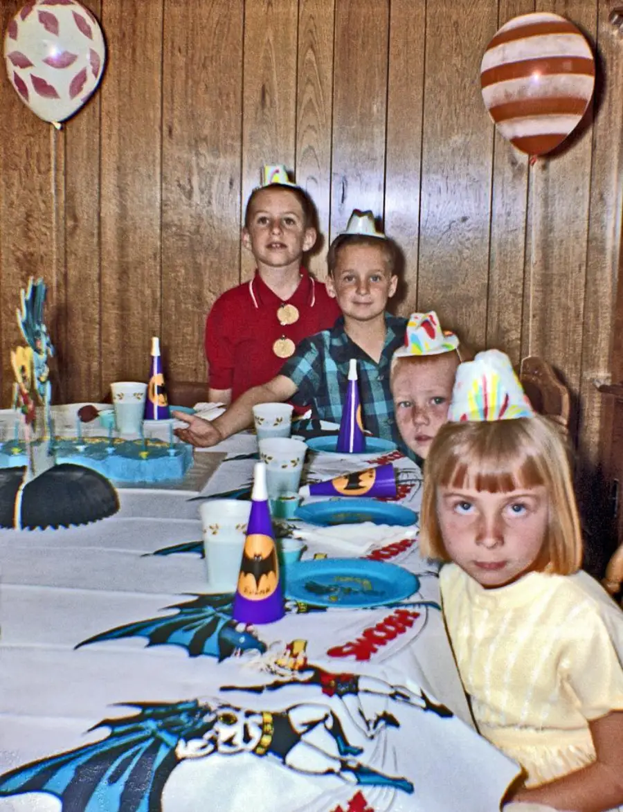 Four children wearing party hats sit at a table decorated with superhero-themed items, including Batman party hats and a Superman tablecloth, with balloons and wood-paneled walls in the background.