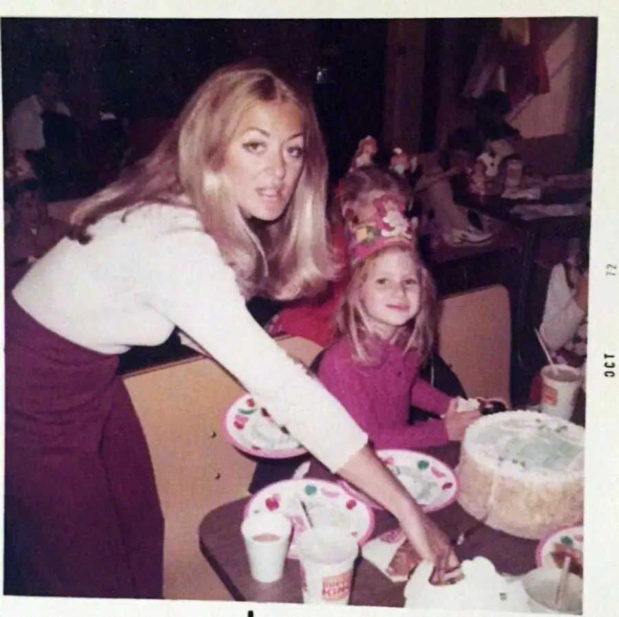 A woman serves cake to a smiling girl wearing a birthday hat at a table decorated with paper plates and cups, surrounded by other children at a party.