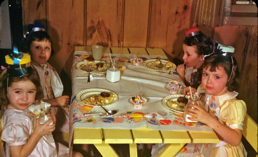 Four young children wearing party hats sit around a small table with plates of food and milk in glass jars, appearing to be at a birthday party. The table is covered with a colorful tablecloth.