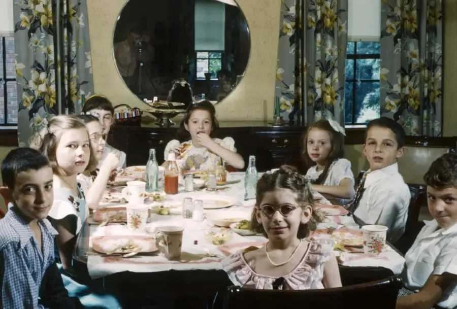A group of children in vintage clothing sit around a table set for a meal, looking toward the camera. The room has floral curtains, a large round mirror, and windows letting in daylight.