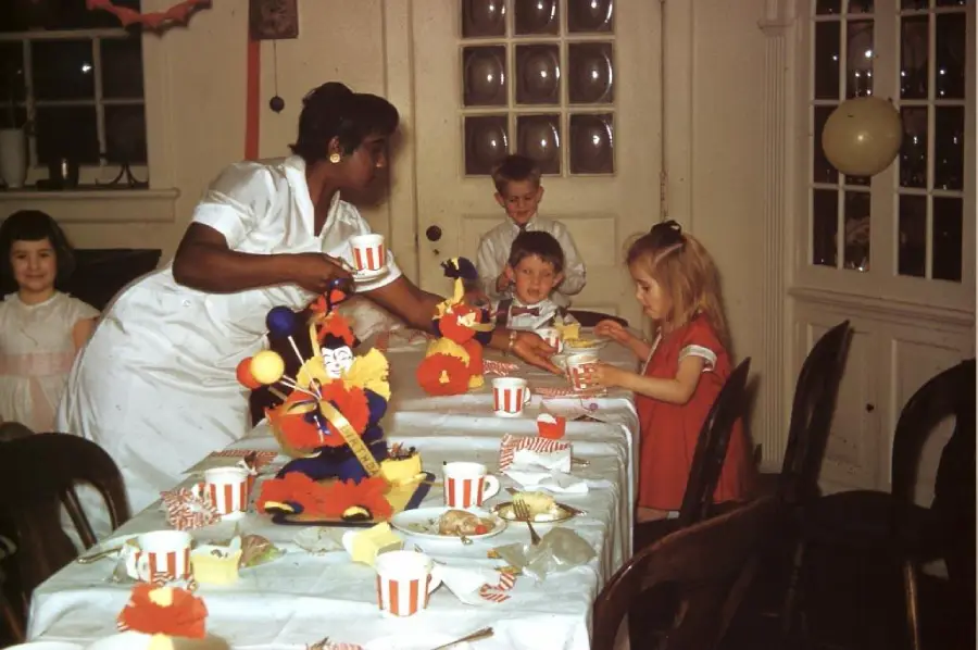 A woman serves drinks to four children at a party table decorated with colorful centerpieces, cups, and party hats in a room with white walls and windows.