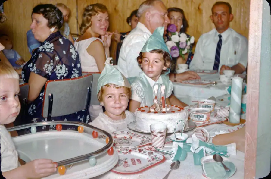 Children wearing party hats sit at a table with a birthday cake, surrounded by adults in a festive indoor setting. The scene is lively with plates, cups, and decorations on the table.