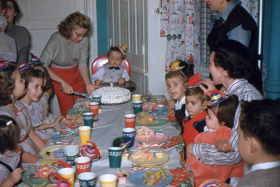 Children wearing party hats sit around a table with colorful cups, plates, and a birthday cake. An adult cuts the cake while others watch, and a baby sits at the head of the table. The room is decorated for a celebration.