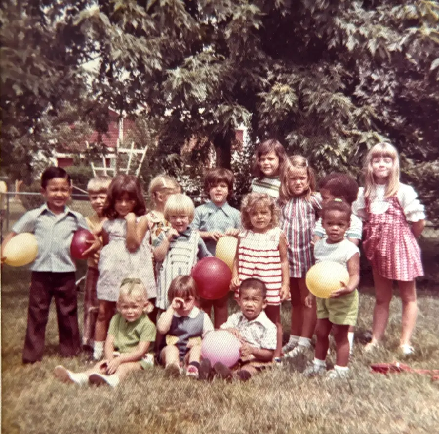 A group of children holding balloons stand and sit on grass in front of leafy trees, smiling and posing for a photo on a sunny day.