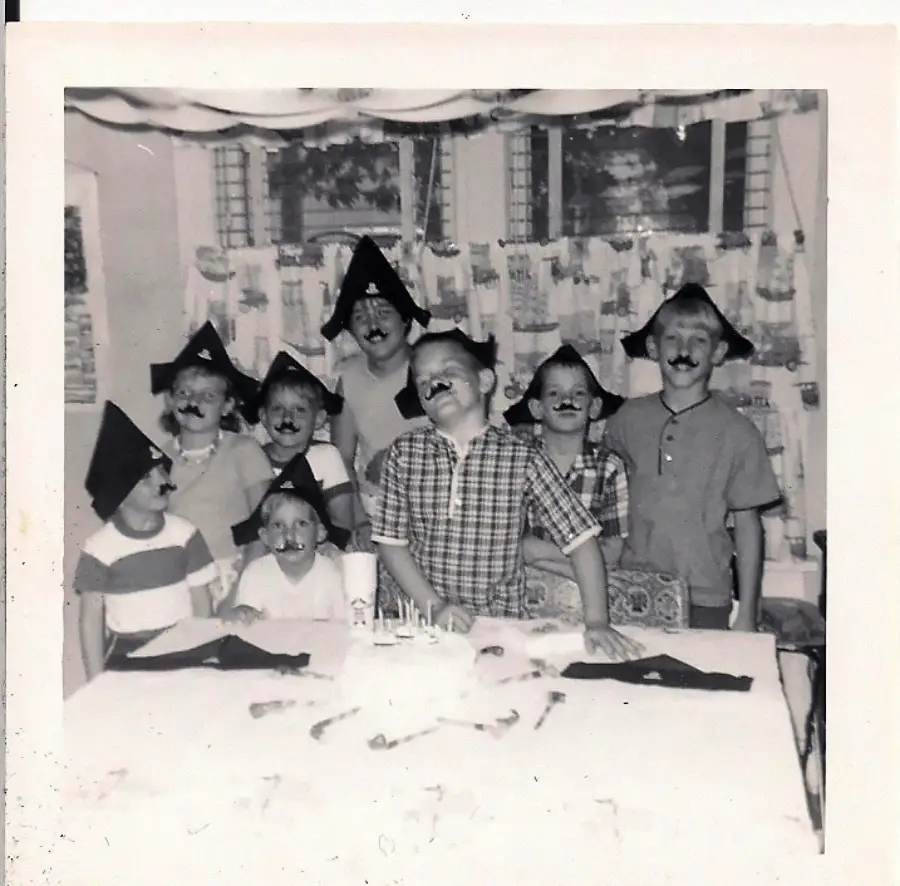 Seven children wearing pirate hats and fake mustaches pose around a table with a birthday cake. The room has decorated windows and a festive atmosphere. The photo is black and white, giving it a vintage feel.
