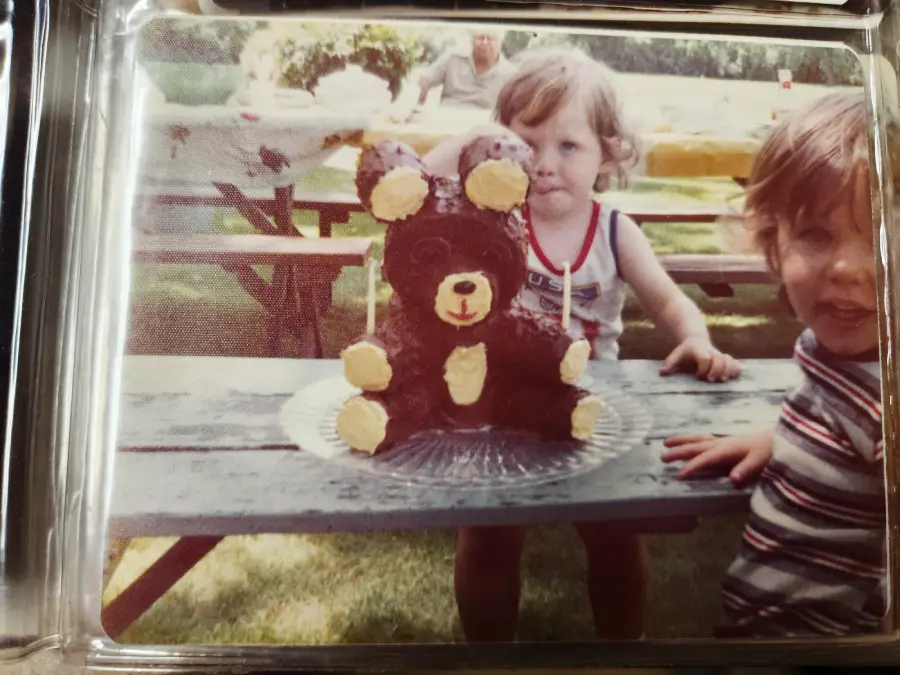 Two young children stand by a picnic table with a chocolate bear-shaped cake on a clear platter outdoors, with trees and an adult in the blurred background.