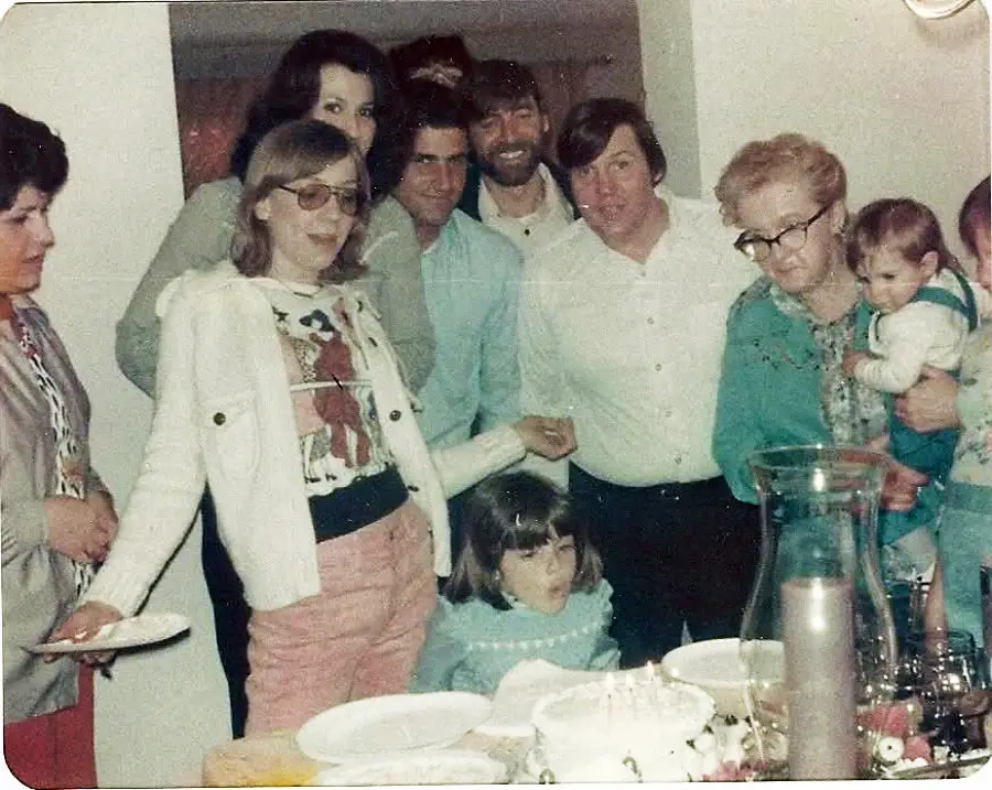 A group of adults and children gathered around a table with a birthday cake, smiling and watching a girl blow out candles.