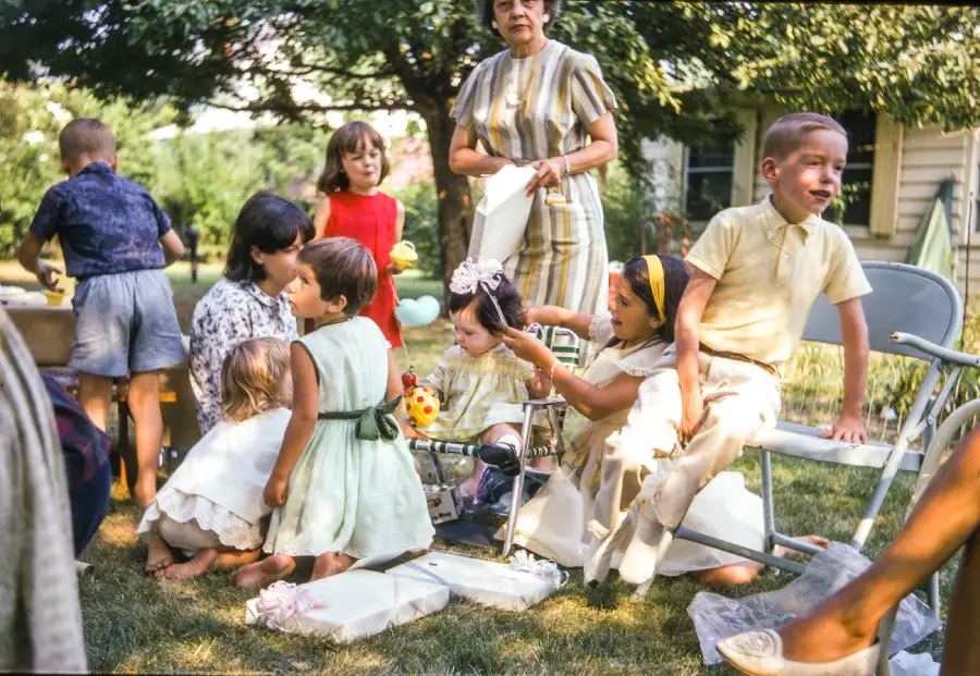 A group of children and a woman gather outdoors at a party. Some kids sit on chairs, others play on the grass. The woman stands in the background, while trees and a house are visible behind them.