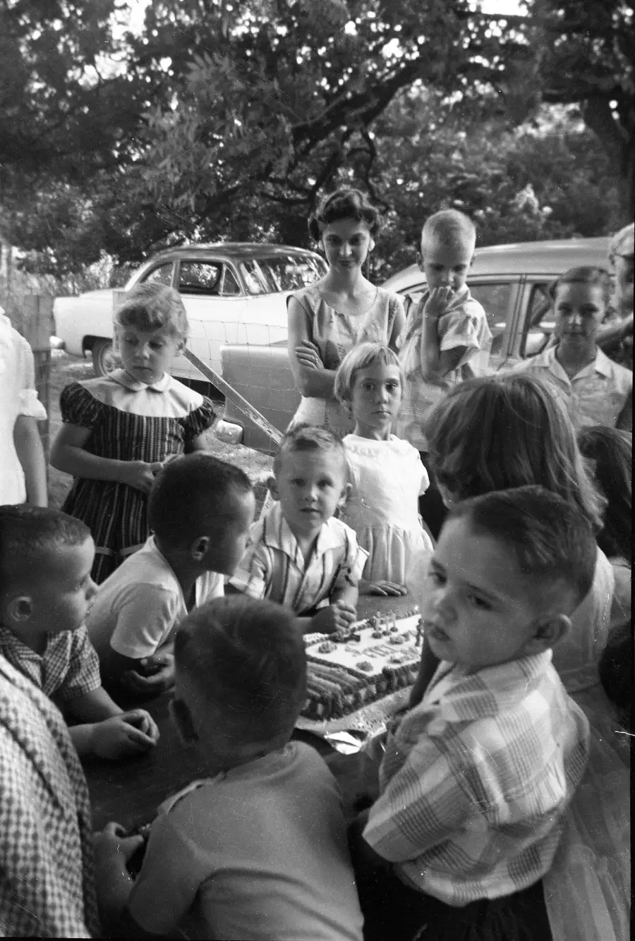 Children gather around a table outdoors with a birthday cake, while adults and cars are seen in the background, capturing a candid vintage party scene.