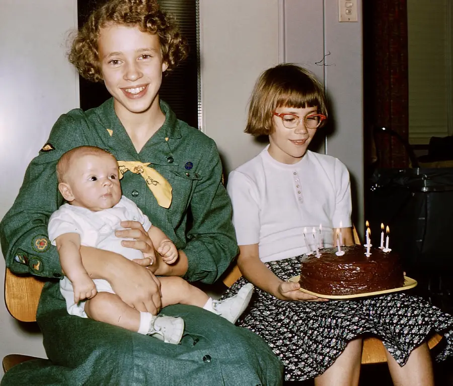 A smiling girl in a green dress holds a baby, while another girl with glasses and a short bob haircut holds a chocolate cake with five lit candles.