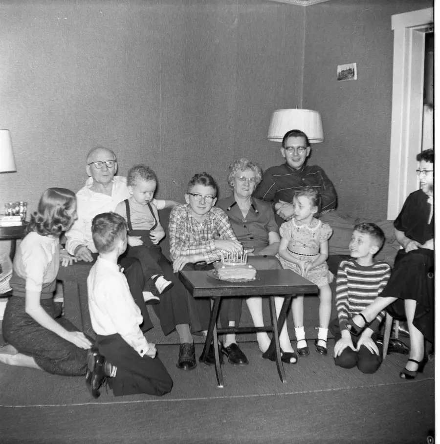 A group of adults and children gather around a table with a birthday cake in a living room, smiling for a photo. The scene appears to be from the mid-20th century.