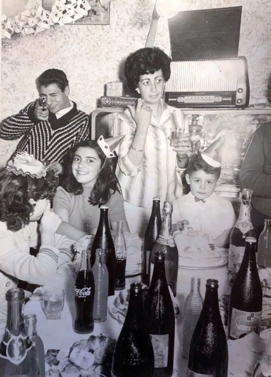 A black-and-white photo of children and adults at a party table with soda bottles. One child wears a party hat, and an adult holds a drink. Another person uses a toy gun. Everyone appears to be enjoying the celebration.
